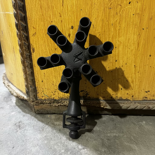 A vintage cast iron multi-spoke fire hydrant cover or decorative fixture, featuring eight cylindrical openings and a central cross-shaped hub, photographed against a yellow wooden panel background.