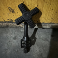 A heavy-duty cast iron cross handle with a textured grip and a long mounting stem, shown resting on a dark concrete floor against a yellow wooden wall.