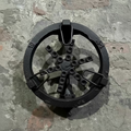 A circular metal wheel-like mechanical part with multiple bolt holes and rusty metal loops, photographed from above on a rough concrete or stone surface.